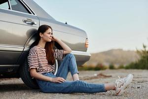 un joven mujer se sienta en el suelo cerca su coche en el lado de el la carretera y mira a el puesta de sol foto