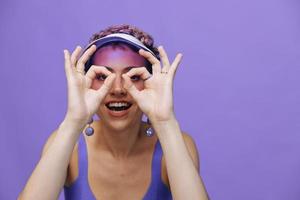 Portrait of a sporty fashion woman posing smiling with teeth at the camera with stylish earrings in her ears in a purple yoga tracksuit and a transparent cap on a purple monochrome background photo