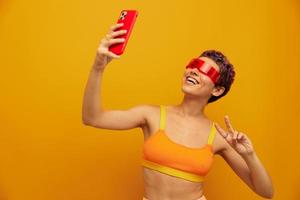 Woman blogger smiling in unusual millennial glasses taking selfies on her phone in sportswear against an orange studio backdrop, free space photo