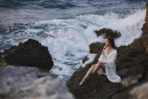 hermosa mujer en un blanco Boda vestir se sienta en el piedras por el Oceano pensativo Mira foto