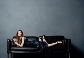 atractivo mujer en negro vestir mentiras en un cuero sofá adentro con elevado piernas foto