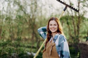 un mujer sonriente hermosamente y mirando a el cámara, un granjero en trabajo ropa y un delantal trabajando al aire libre en naturaleza y participación un rastrillo a reunir césped y forraje para el animales en el jardín foto