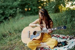 Girl hippie woman playing guitar in eco-friendly clothes sitting on the ground outside in nature in the fall watching the sunset photo