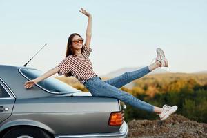un mujer con un coche detenido en el la carretera a descanso en el viaje elevado su brazos y piernas desde felicidad y un hermosa paisaje foto