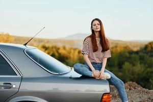 A woman car driver sits on the trunk of a car and looks into the distance admiring a beautiful view of autumn nature and mountains photo