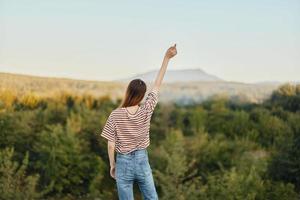 A young woman stands with her back to the camera with her hands up in a T-shirt and jeans in nature and enjoys a beautiful view of the mountains. Autumn travel to nature lifestyle photo