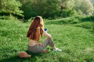 Top view of a woman in an orange top and green pants sitting on the summer green grass with her back to the camera with her phone, a young freelance student's concept of work and leisure photo