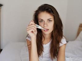 woman in white t-shirt sits on bed thermometer in mouth temperature check photo