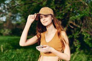 mujer blogger con teléfono en mano en naturaleza en contra un fondo de verdor sonriente en el Brillo Solar vistiendo un gorra después hacer ejercicio foto