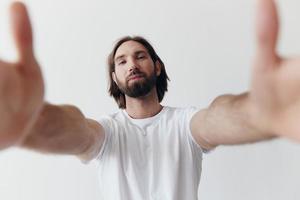 Happy adult man with a beard smiles and pulls the cook into the camera listening to music in headphones in a distressed t-shirt on a white isolated background photo