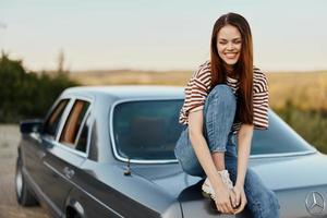 A young woman sits on the trunk of a car, laughs and rests after a difficult road and admires nature with a beautiful view. Stopping is also part of the journey photo