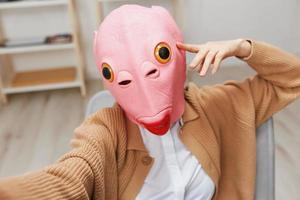 gracioso rubia dama en rosado pescado máscara calentar suéter haciendo selfie utilizar teléfono sentado en Sillón a moderno hogar interior. pausa desde trabajar, tomar un romper, social medios de comunicación en gratis hora concepto. amplio ángulo foto
