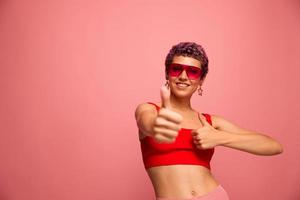 Fashion portrait of a woman with a short haircut in colored sunglasses with unusual accessories with earrings smiling on a pink bright background showing thumbs up at the camera photo