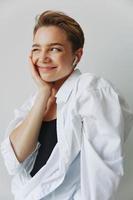 Young woman teenager listening to music with infertile headphones and dancing home, grinning with teeth with a short haircut in a white shirt on a white background. Girl natural poses with no filters photo