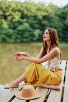A hippie woman sits with her hat on a bridge by a lake with her hands up in the air on a nature trip and smiling happily in eco-clothing. Relaxed lifestyle photo