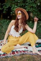 Hippie woman smiling in eco clothing yellow pants, white knit top, hat and yellow glasses sitting on plaid in park watching sunset, lifestyle camping trip photo