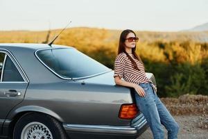 elegante mujer en a rayas camiseta Gafas de sol y pantalones cerca gris coche en naturaleza foto