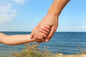 Man holds hand of child on background of sea and sky Concept of love, care, friendship, trust in family. photo