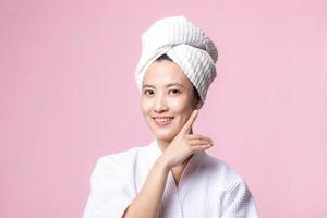 hermosa joven asiático mujer contento con limpiar cara piel en toalla y bata de baño, spa traje en rosado antecedentes. protección de la piel, tratamiento, bienestar terapia, facial cuidado, belleza hembra salud, cosmetología concepto foto