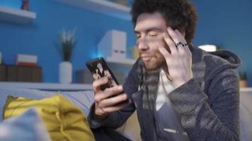 Jeune homme mensonge sur le canapé dépense une lot de temps avec le sien téléphone intelligent, obtient intoxiqué à il. téléphone intoxiqué Jeune homme mensonge sur canapé à maison, à la recherche à cellule téléphone, surfant social médias, SMS. video