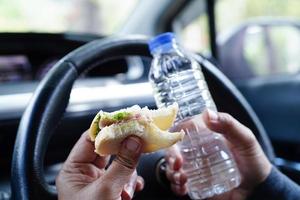 Asian woman driver hold cold water and sandwich bread for eat and drink in car, dangerous and risk an accident. photo