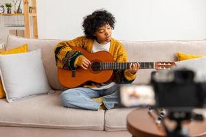 Blogger guitarist. African american girl blogger playing guitar talking to webcam recording vlog. Social media influencer woman streaming at home indoors. Music content creator broadcast tutorial. photo
