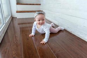 Little crawling baby girl one year old siting on floor in bright light living room near window smiling and laughing photo