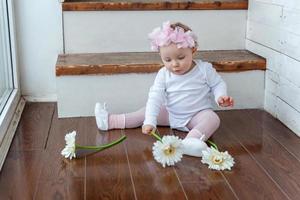 pequeño bebé niña vistiendo primavera guirnalda emplazamiento en piso en brillante ligero vivo habitación cerca ventana y jugando con gerbera flores foto