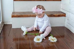 pequeño bebé niña vistiendo primavera guirnalda emplazamiento en piso en brillante ligero vivo habitación cerca ventana y jugando con gerbera flores foto