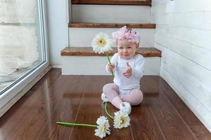 Little baby girl wearing spring wreath siting on floor in bright light living room near window and playing with gerbera flowers photo