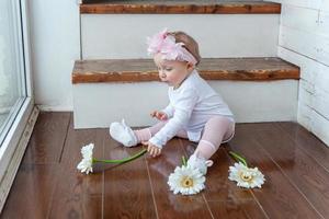 pequeño bebé niña vistiendo primavera guirnalda emplazamiento en piso en brillante ligero vivo habitación cerca ventana y jugando con gerbera flores foto