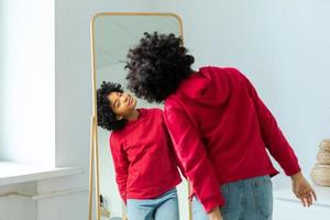 amor tú mismo. hermosa joven sonriente africano americano mujer bailando disfrutando su espejo reflexión. negro dama mirando a espejo mirando confidente y contento. yo amor concepto. foto