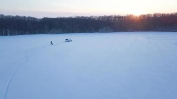 View from height to man is riding a snowboard on a snow-covered field on a cable behind a car video