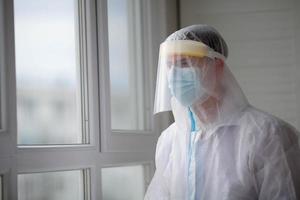 A sad tired doctor in a protective suit and a medical mask sits near a window in a hospital. Pandemic, covid - 19. photo