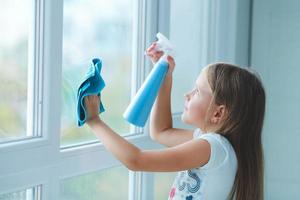 A little girl sprinkles water from a bottle on the window and wipes it off with a rag. The child helps with cleaning the house. photo