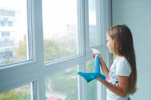 A little girl sprinkles water from a bottle on the window and wipes it off with a rag. The child helps with cleaning the house. photo