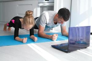 padre y hija son hacer ejercicio, haciendo el tablón mientras mirando a el ordenador portátil. el familia obras de teatro Deportes en línea a hogar en el cocina. familia obras de teatro Deportes durante el covid-19 pandemia foto