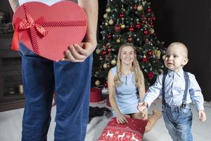 Navidad sorpresa. un hombre sostiene un rojo regalo caja en el antecedentes de su esposa y niño. familia en nuevo años vacaciones. el pequeño hijo carreras a recibir un Navidad regalo. foto