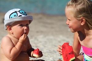 Portrait of children of sister and brother on the beach with watermelon. Girl and little boy are eating on the beach. The children had a picnic by the sea. photo