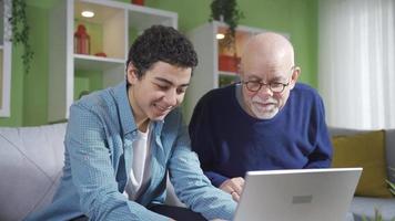 curioso abuelo mirando a qué su linda nieto es haciendo mientras mirando a ordenador portátil. nieto trabajando en ordenador portátil en sofá y abuelo acecho él con curioso ojos. video
