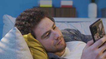 Young man lying on sofa at home, looking at phone, happy. Happy young man lying on sofa at home, mind looking at mobile phone, browsing social media, texting. Phone Addicted Teen. video