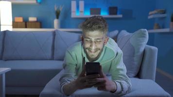 knap blond jong Mens gebruik makend van telefoon is gelukkig en prettig. jong Mens aan het liegen Aan sofa alleen Bij huis gebruik makend van telefoon en glimlachen gelukkig en speels video