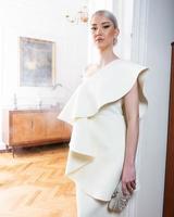 A woman in a white dress stands in a room with a wooden cabinet and a plant in the background. photo