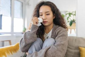 Unhappy young Asian woman crying alone close up, depressed girl sitting on couch at home, health problem or thinking about bad relationships, break up with boyfriend, divorce photo