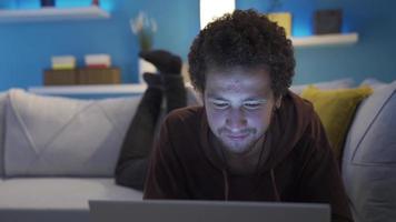 a homem deitado baixa e olhando às a computador portátil é sorridente e dentro Boa espíritos. feliz jovem homem deitado em sofá olhando às computador e sorridente. video