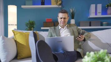 Casual dressed carefree man using smartphone on sofa at home, looking at laptop. video