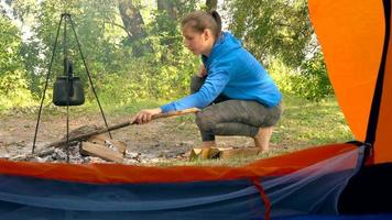 Aussicht von ein Tourist Zelt - - ein Frau setzt Brennholz auf ein Feuer auf welche das Kessel ist erhitzt video