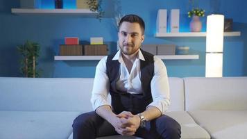 Charismatic and attractive businessman sitting on sofa at home smiling at camera and looking around. video
