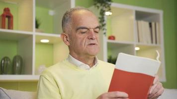 Old man reading a thoughtful and dreamy book feeling calm and relaxed. Mature man reading a book at home. Knowledge and culture concept. video