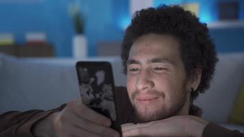 Happy and sympathetic young man looking at his smartphone at home, feeling affection, smiling. Sympathetic cute young man looking at smart mobile phone at home smiling and being happy. video